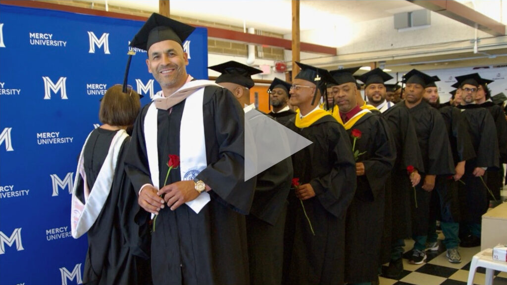 Hudson Link Graduates standing in line wearing caps and gowns. Play button overlays image to indicate it is clickable (for seeing the video).